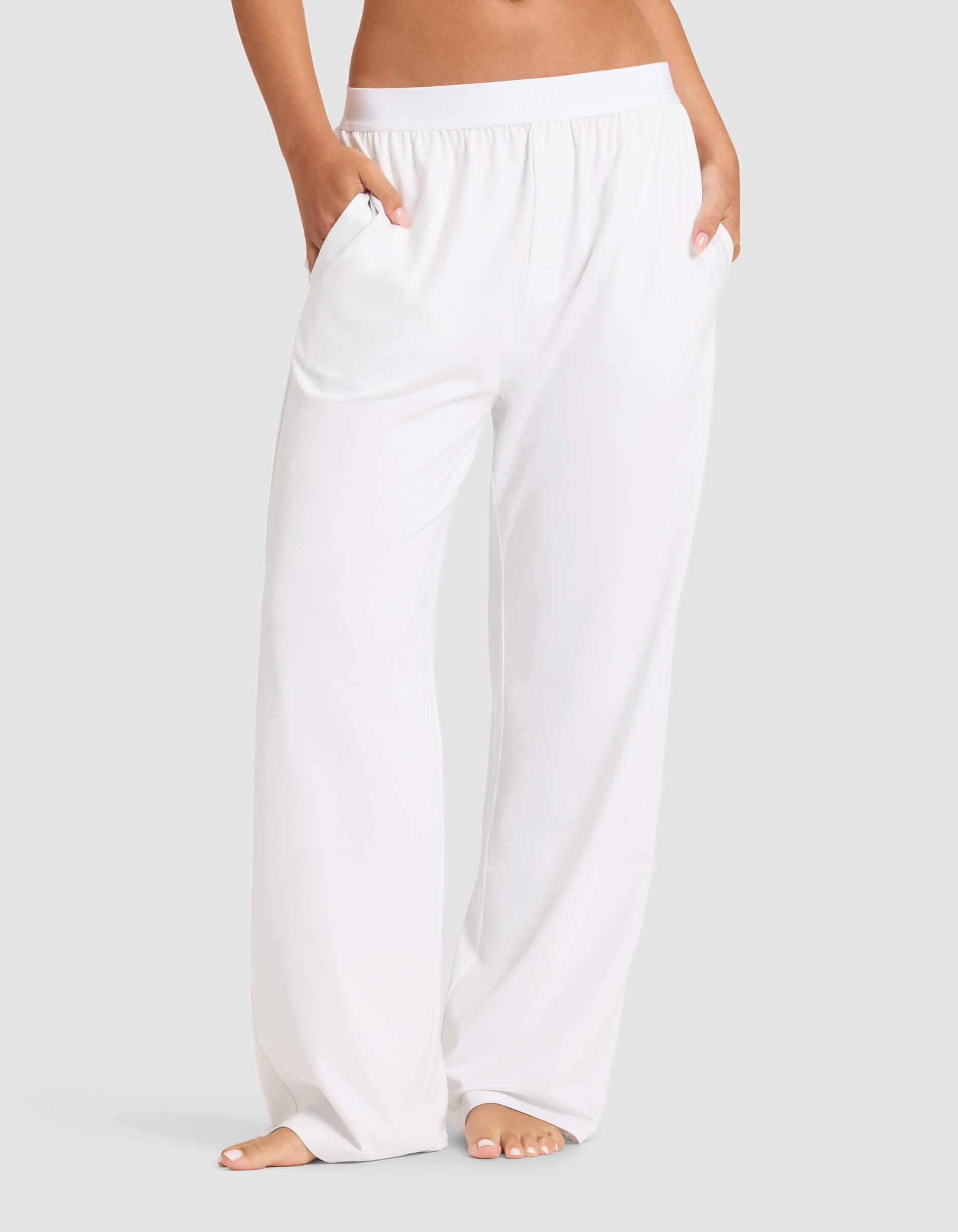 A woman models Cozy Earth’s Women's First Layer Pajama Pant—loose-fitting, white with an elastic waistband and pockets—standing barefoot against a light gray background.|Color:White