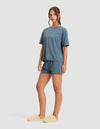 A woman models the Cozy Earth Women's First Layer Pajama Short Sleeve Tee & Boxer Short Set in blue, paired with beige fuzzy slippers, against a plain light background.|Color:Dawn