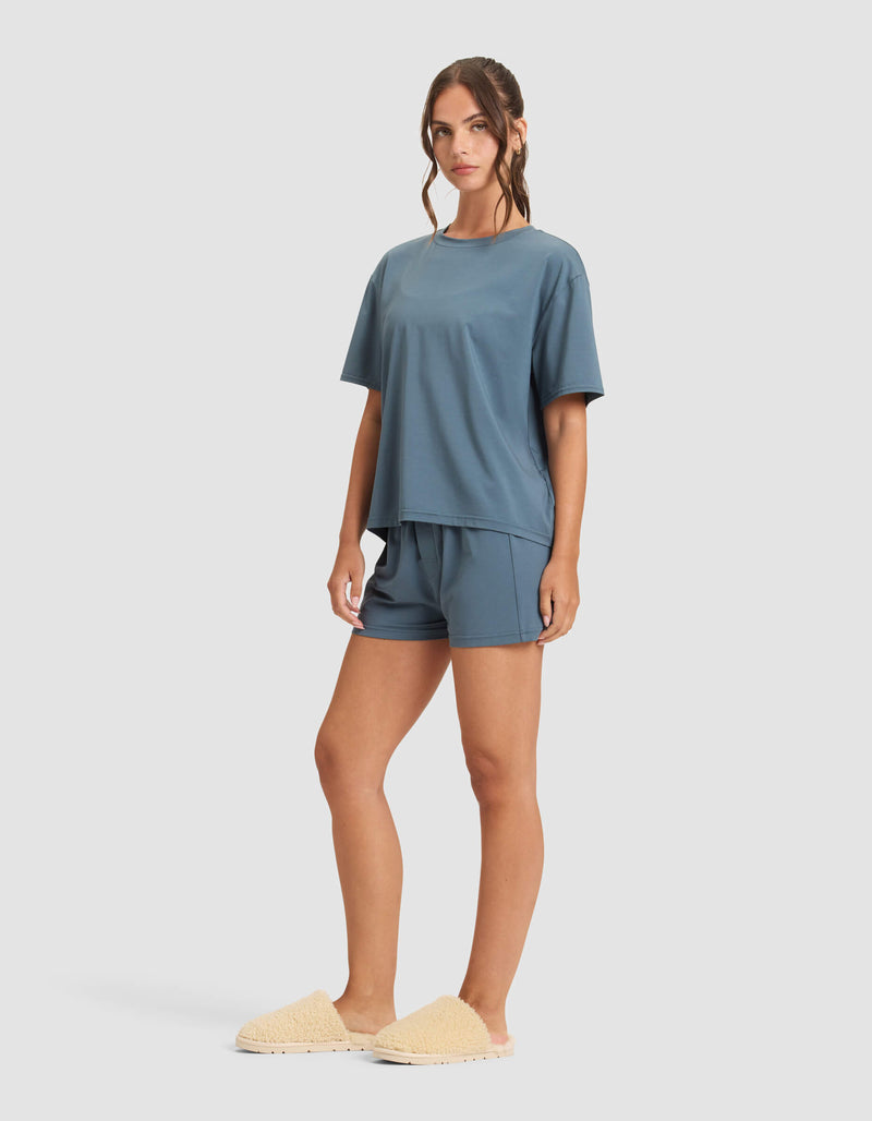 A woman models the Cozy Earth Women's First Layer Pajama Short Sleeve Tee & Boxer Short Set in blue, paired with beige fuzzy slippers, against a plain light background.