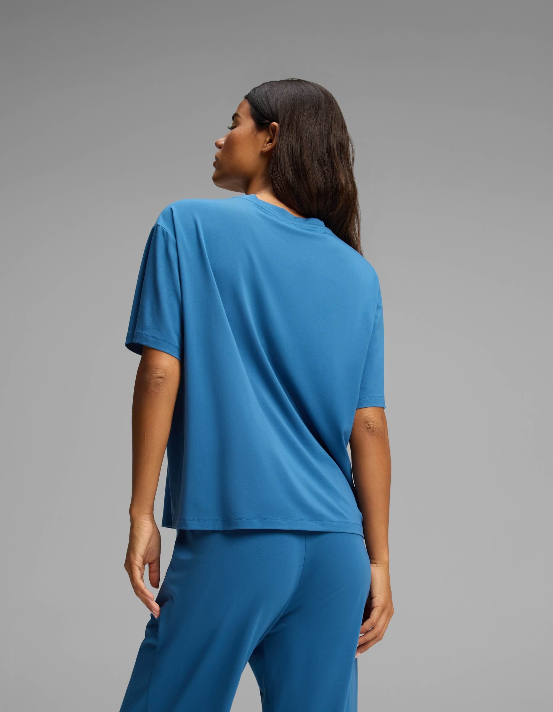 A person with long dark hair is seen from behind, wearing the Cozy Earth Women’s First Layer Pajama Short Sleeve Tee in blue along with matching blue pants, standing against a plain gray background.