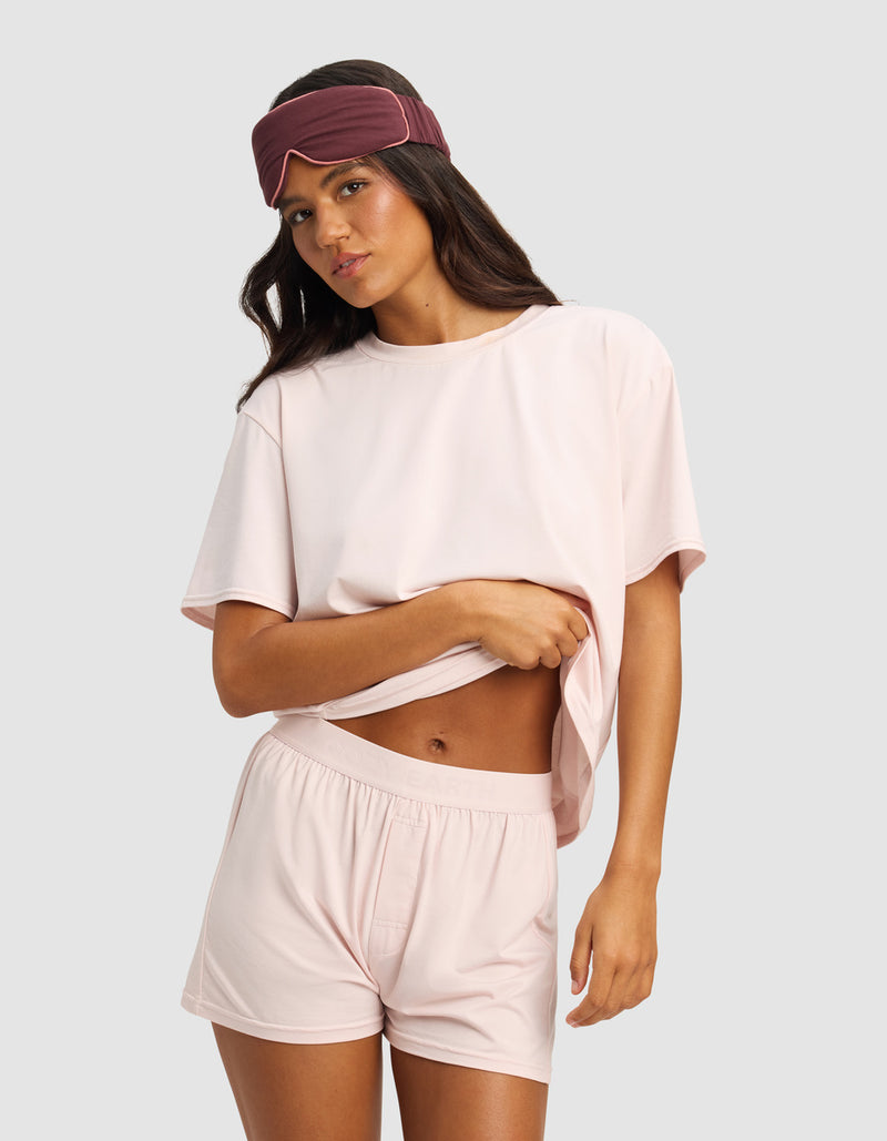 A woman stands against a light gray background wearing the Cozy Earth Women's First Layer Pajama Short Sleeve Tee & Boxer Short Set in light pink, paired with a dark mauve sleep mask and lifting part of her shirt with one hand.