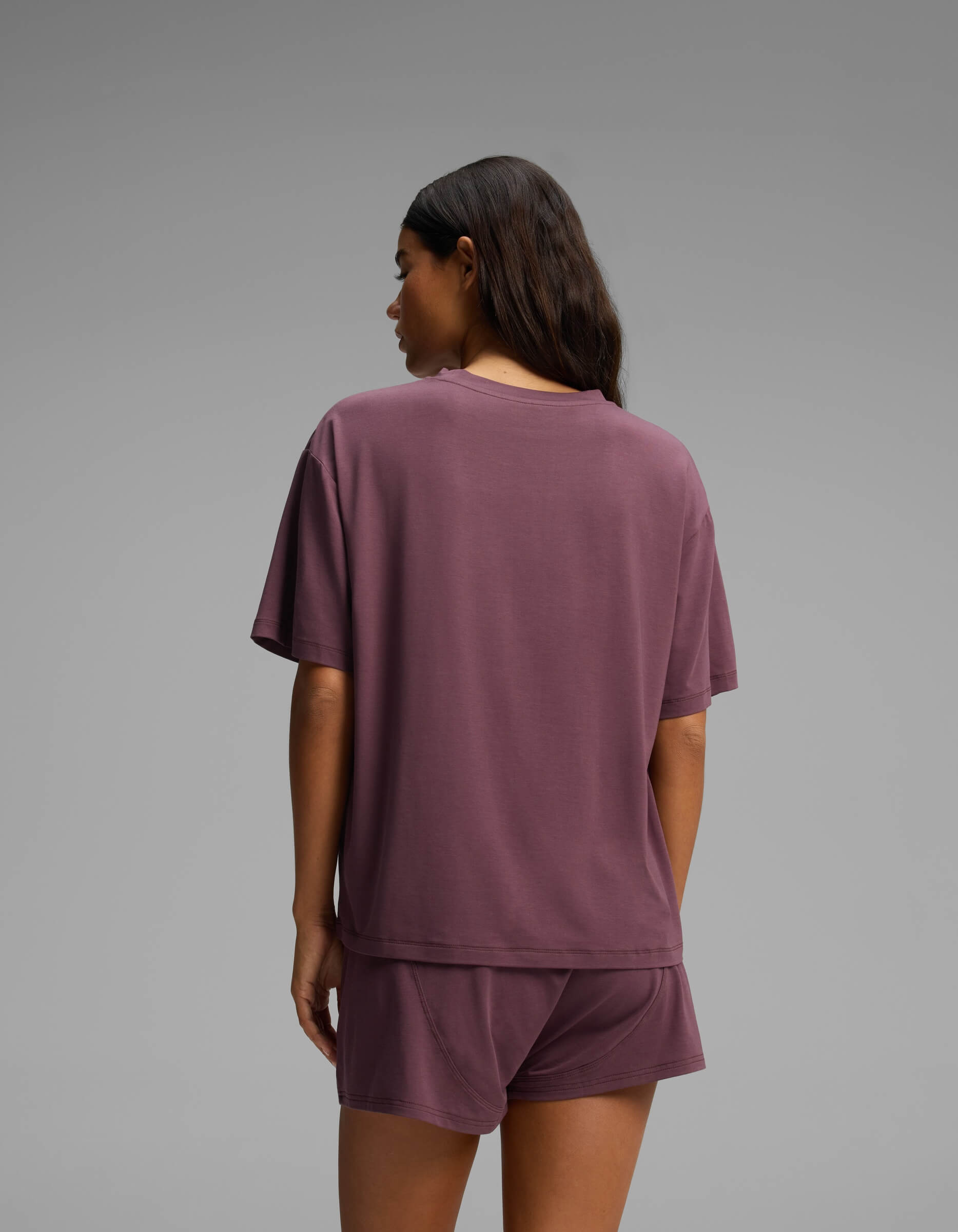 A person with long dark hair stands with their back to the camera against a plain gray background, wearing Cozy Earth’s Women’s First Layer Pajama Short Sleeve Tee and matching shorts in mauve.