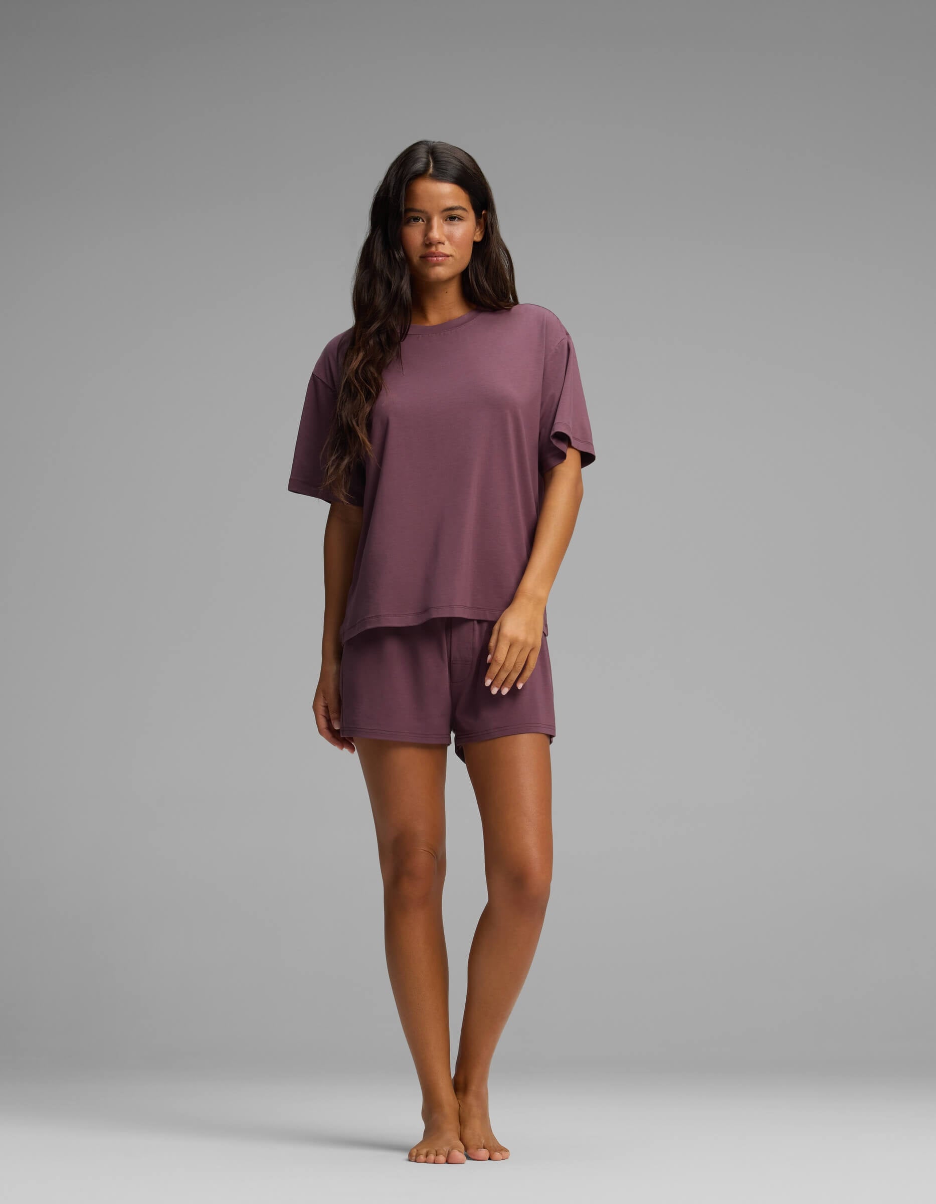 A woman stands barefoot against a plain gray background, wearing the Cozy Earth Women’s First Layer Pajama Short Sleeve Tee and matching purple shorts.