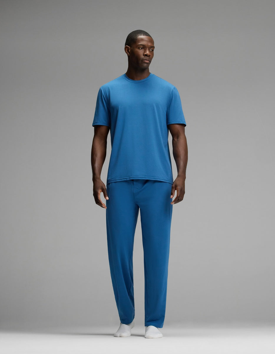A man stands against a gray background wearing the Cozy Earth Men’s First Layer Pajama Short Sleeve Tee & Pant Set in blue with white socks, looking slightly to the side.|Color:Anchor