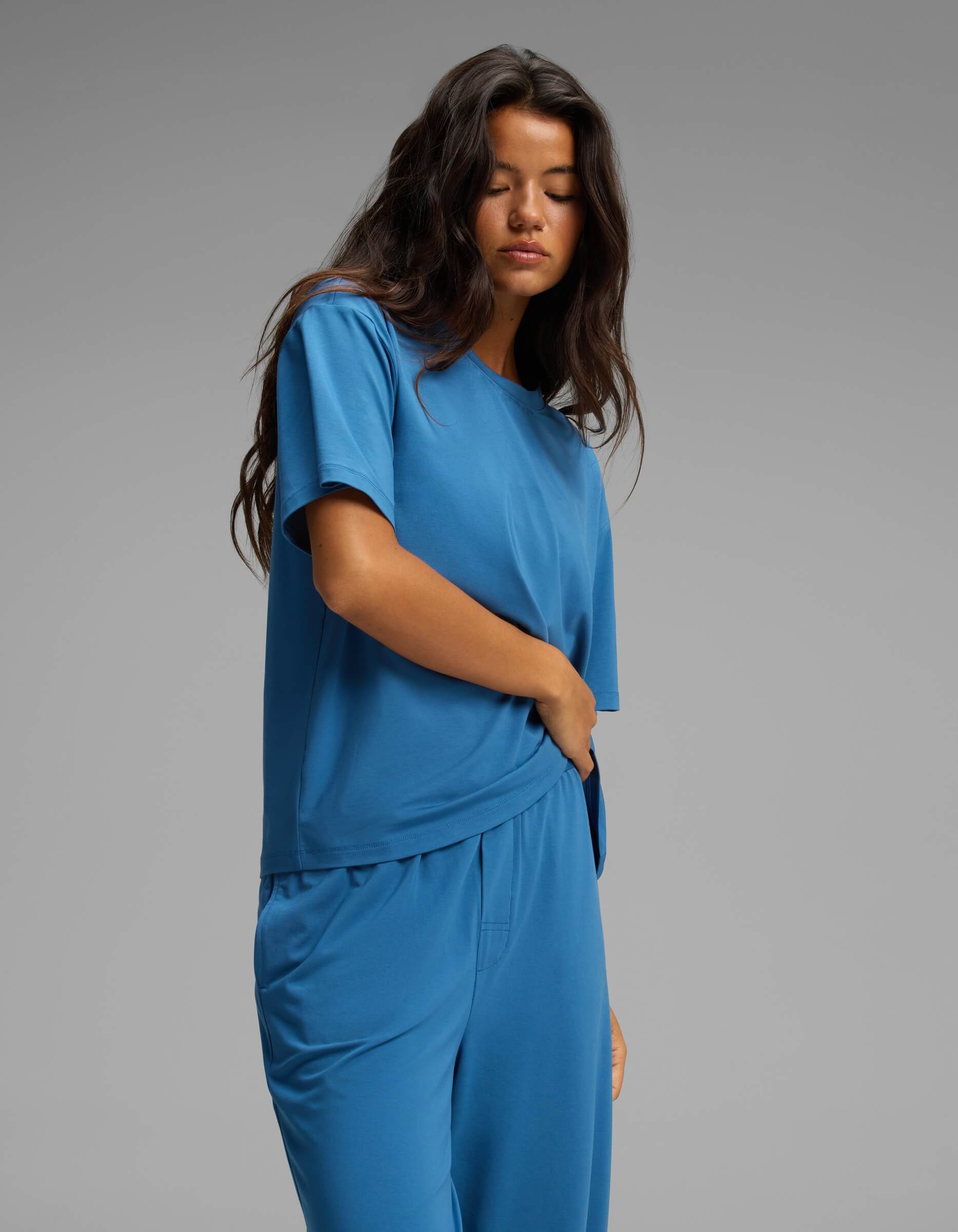 A woman with long dark hair wears the Cozy Earth Women’s First Layer Pajama Short Sleeve Tee & Pant Set in blue, standing against a plain gray background with her eyes closed and one hand resting on her waist.