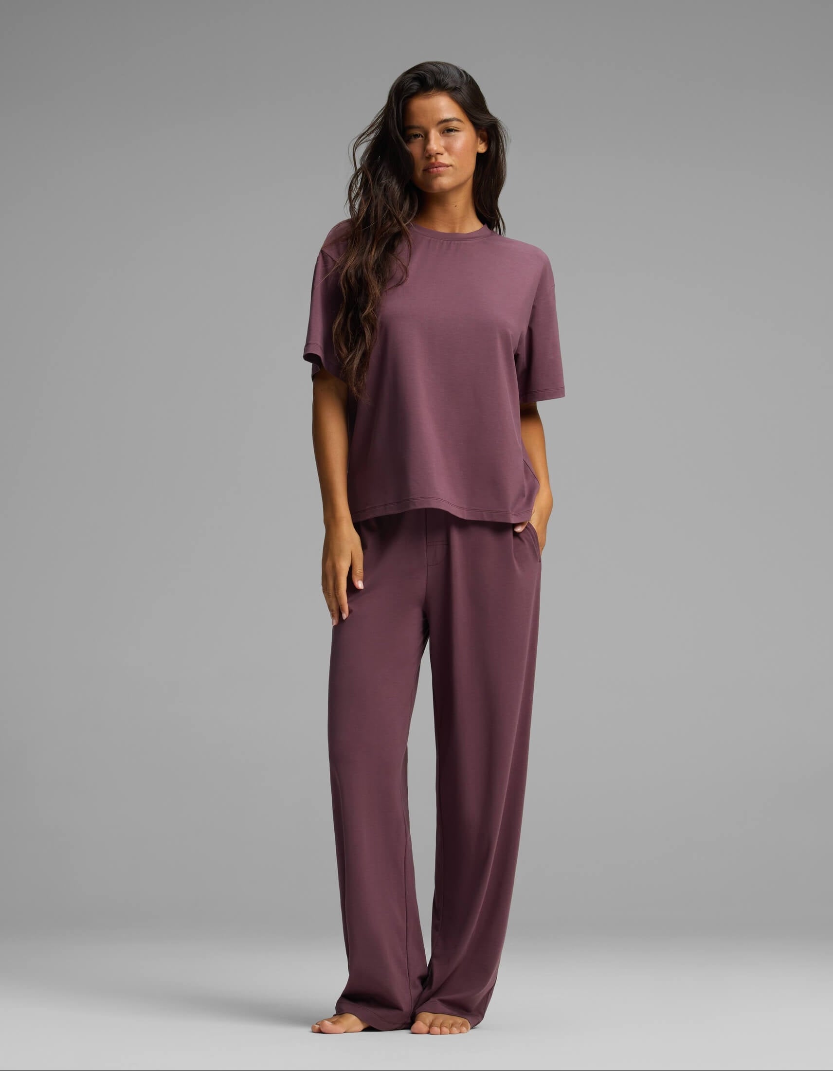 A woman with long dark hair is seen standing barefoot against a plain gray background, wearing the Cozy Earth Women’s First Layer Pajama Short Sleeve Tee & Pant Set in a loose-fitting purple style with matching wide-leg pants.|Color:Fig