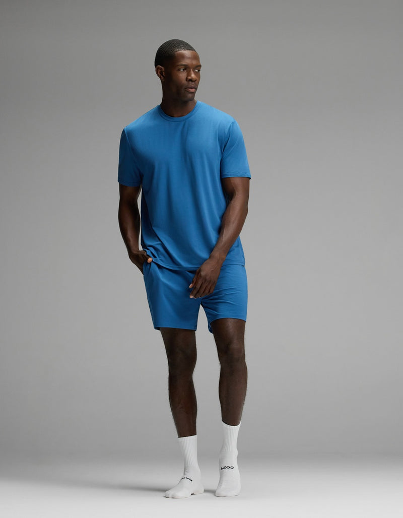 A man stands in a studio wearing the Cozy Earth Men’s First Layer Pajama Short Sleeve Tee & Short Set in blue, paired with white socks. The background is plain and gray, and he gazes off to the side with one hand casually resting in his shorts pocket.
