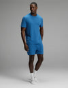 A man stands against a plain gray background wearing the Cozy Earth Men’s First Layer Pajama Short Sleeve Tee & Short Set in blue, paired with white socks. He looks slightly to the side with one hand in his pocket.|Color:Anchor