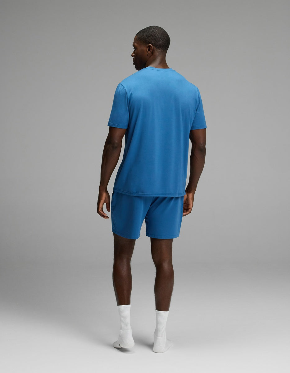 A man, seen from behind, wears the Cozy Earth Men’s First Layer Pajama Short Sleeve Tee & Short Set in blue with white socks. He stands against a plain gray background, showcasing a clean and simple setting.