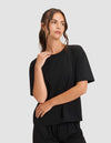 A woman wears the Cozy Earth Women's First Layer Pajama Short Sleeve Tee and matching shorts in black, standing against a light gray background, gazing to the side with one hand near her face and the other on her arm.|Color:Jet Black