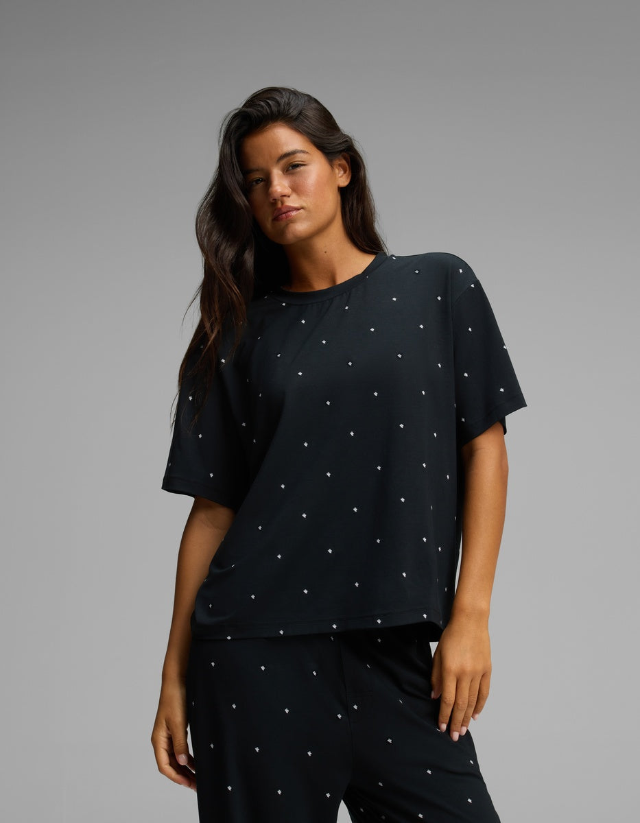 A woman with long dark hair stands against a plain gray background, wearing the Cozy Earth Women’s First Layer Pajama Short Sleeve Tee and matching black pants, both featuring small white polka dots.|Color:Mini Ghost