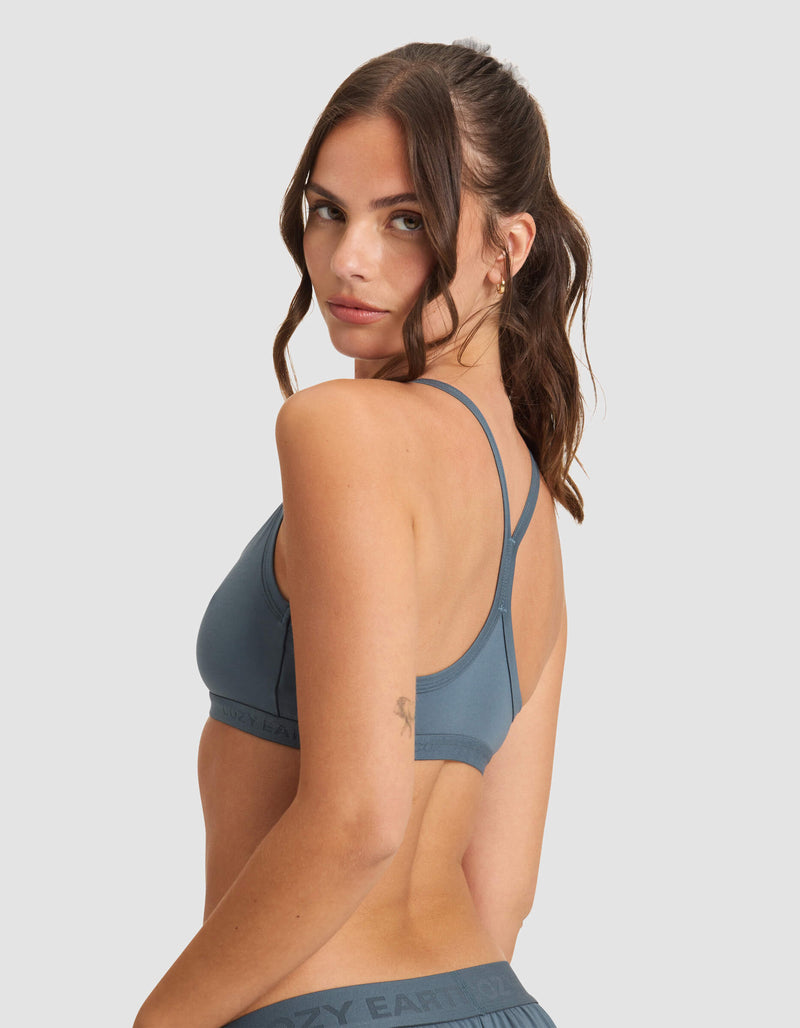 A woman with long brown hair looks over her shoulder, wearing the Cozy Earth Women's First Layer Pajama Soft Bra in blue and matching bottoms against a plain light gray background.