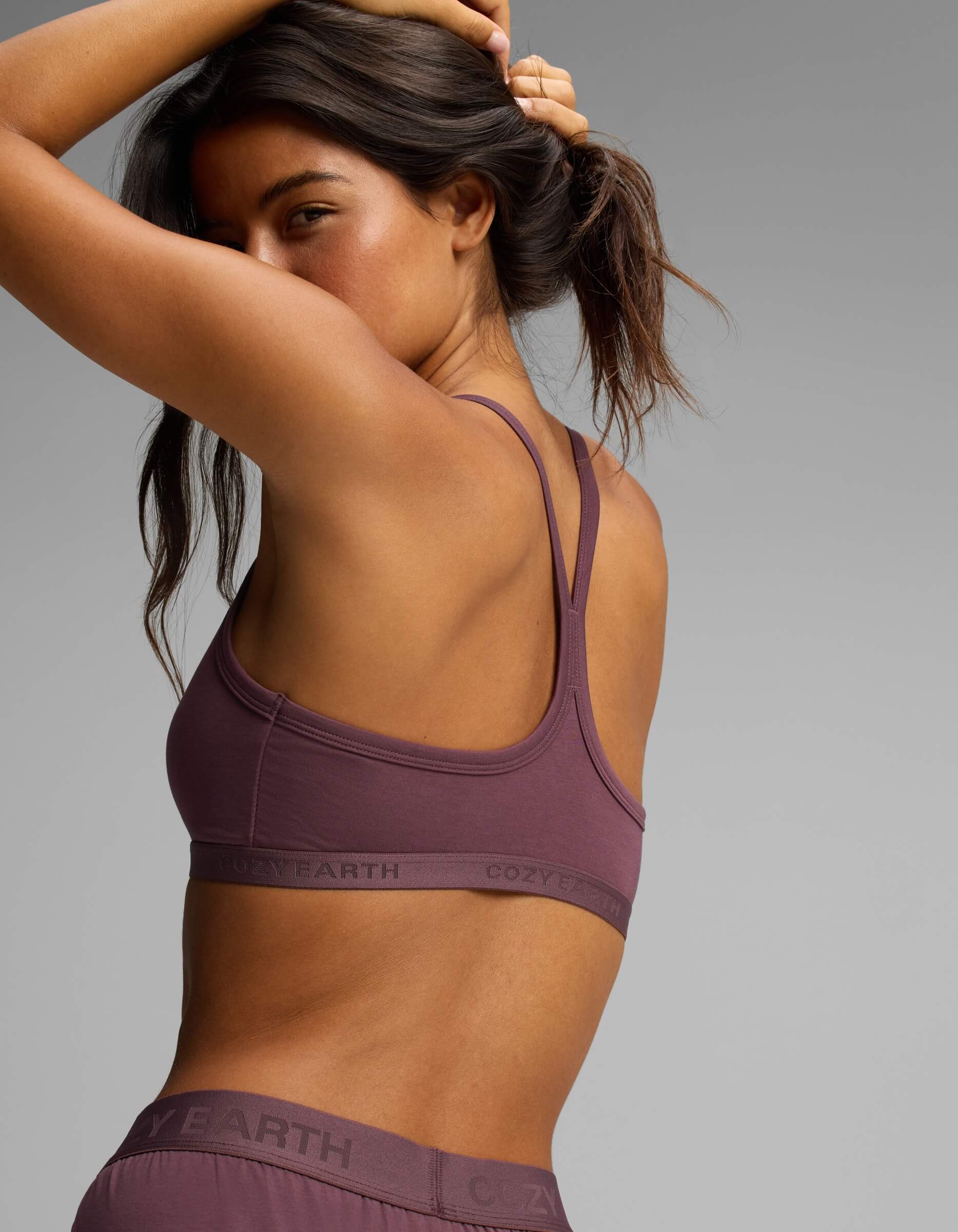 A woman wearing the Cozy Earth Women’s First Layer Soft Bra and matching bottoms, featuring the “Cozy Earth” branding on the band, poses against a plain gray background with one arm raised and looking over her shoulder.