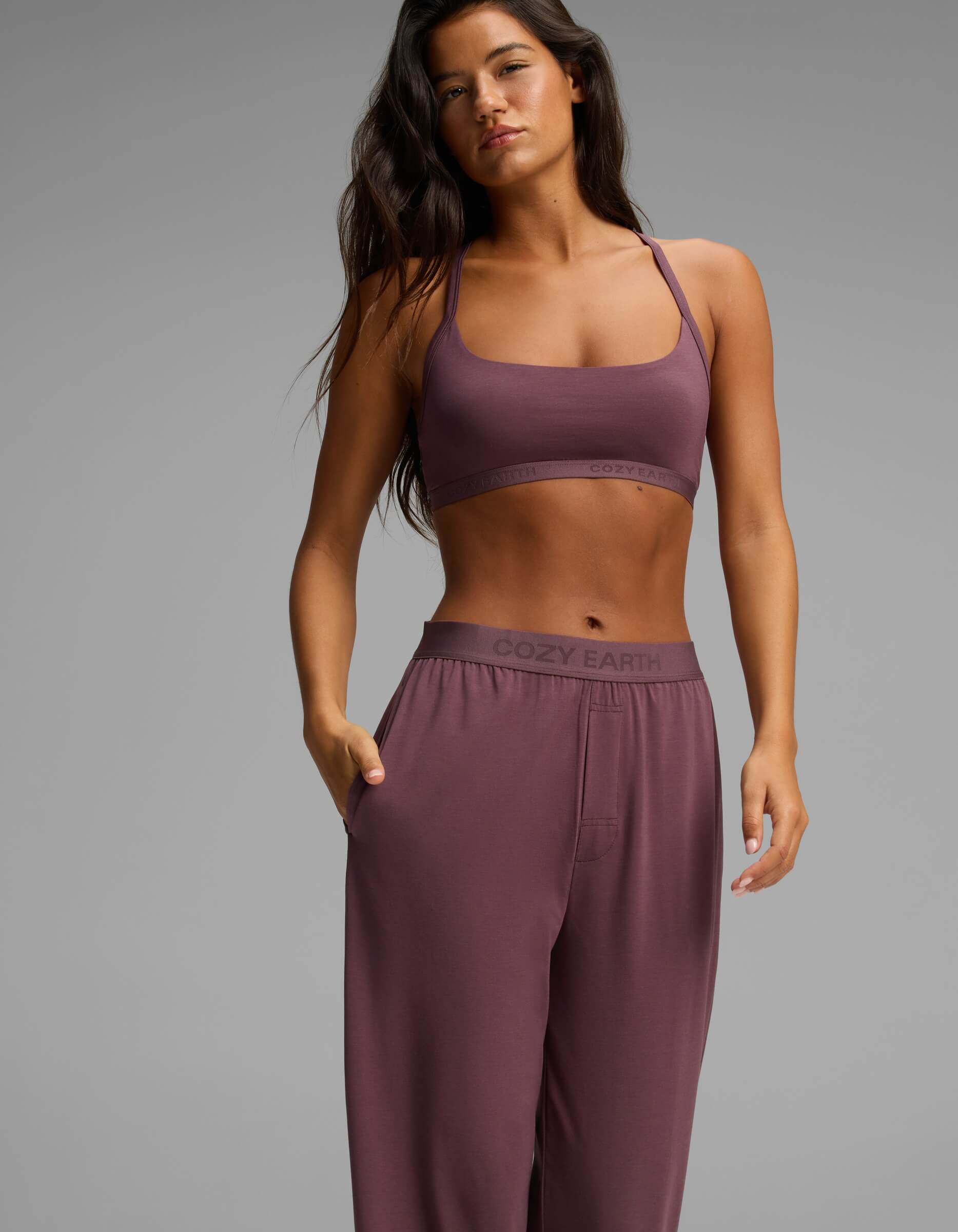 A woman poses against a plain gray background wearing Cozy Earth’s Women’s First Layer Pajama Soft Bra & Pant Set in mauve, featuring a branded sports bra and loose pants.