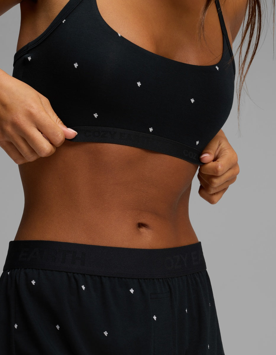 A woman wearing the Cozy Earth Women’s First Layer Soft Bra and matching black shorts with small white patterns adjusts her top, revealing part of her midriff against a neutral background.|Color:Mini Ghost