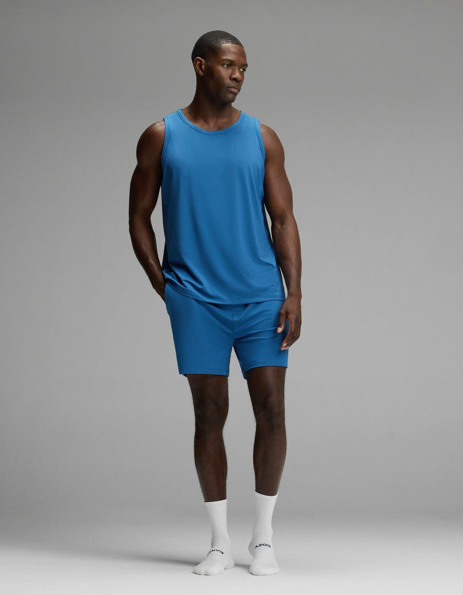 A man stands in a studio wearing the Cozy Earth Men’s First Layer Pajama Tank in blue, paired with matching blue shorts and white socks. He looks slightly to the side with his hands relaxed by his sides.
