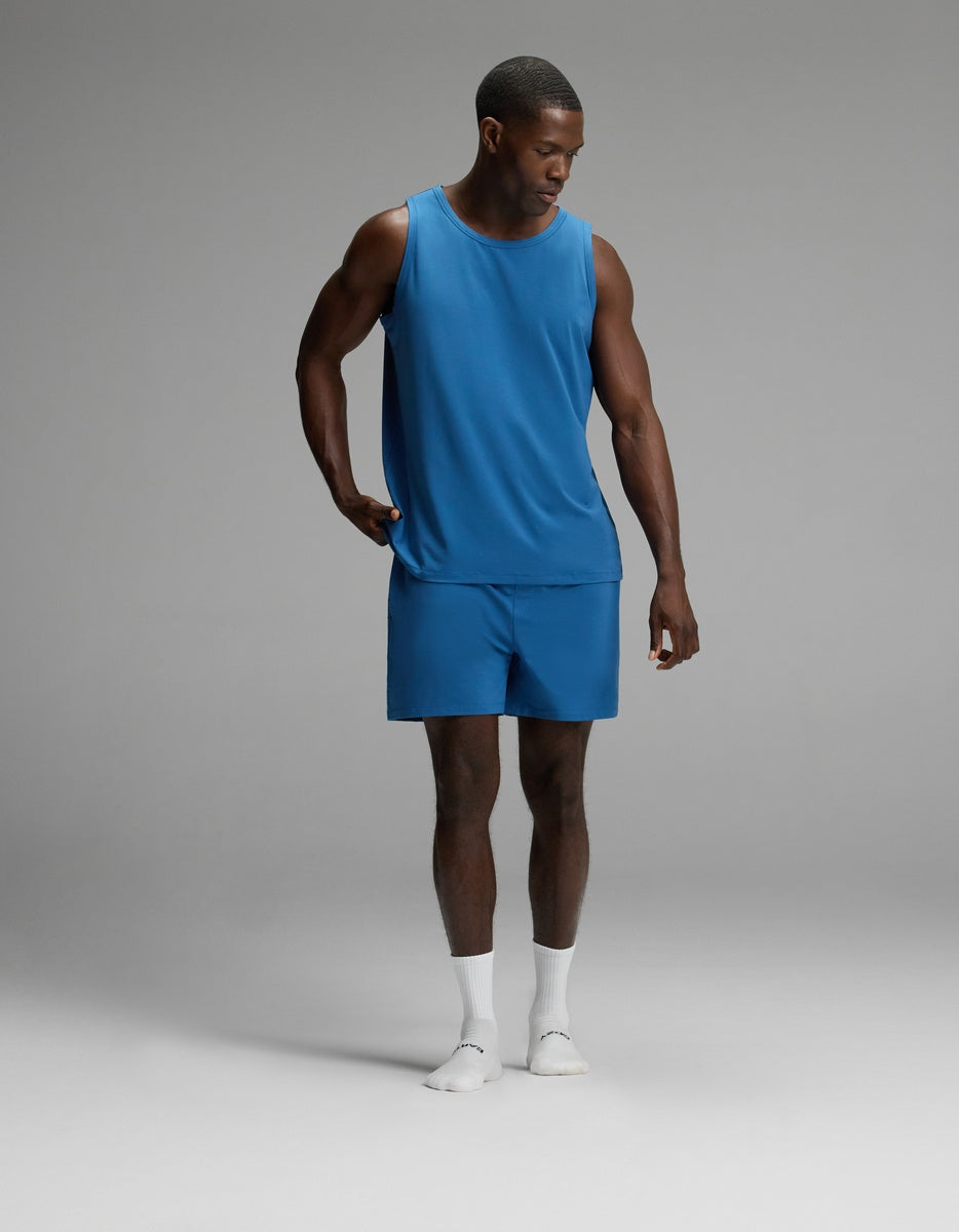 A man stands in a studio wearing the Cozy Earth Men’s First Layer Pajama Tank in blue, paired with matching blue shorts and white socks, looking down with one hand on his hip.