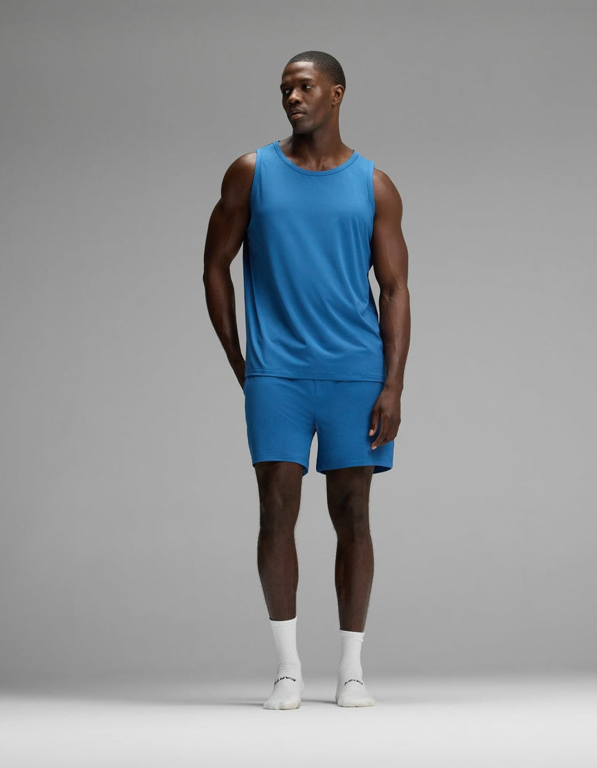 A man stands in a studio against a plain gray background wearing the Cozy Earth Men’s First Layer Pajama Tank & Short Set in blue, paired with white socks and white shoes.
