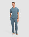 A person stands barefoot against a plain background, wearing Cozy Earth's Men's First Layer Pajama Pant in blue with a matching top. Their left hand is in a pocket, and they face forward with a neutral expression.|Color:Dawn