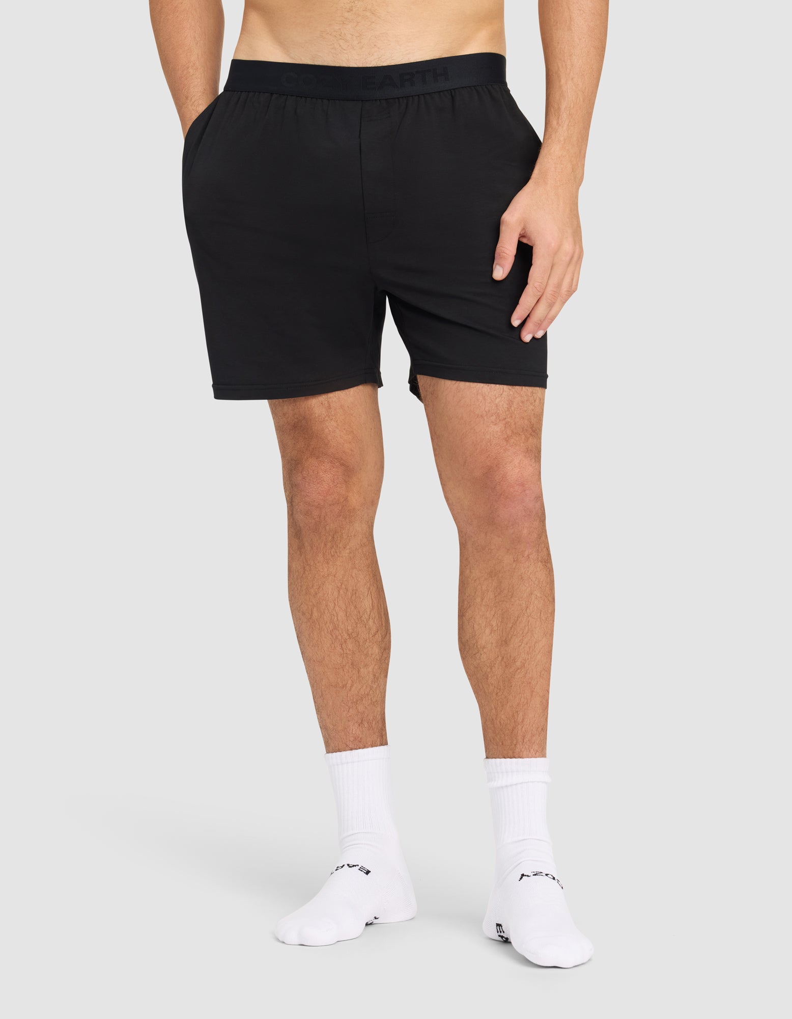 A man models Cozy Earth's Men's First Layer Pajama Short in black with white crew socks, standing on a plain light gray background. The image is cropped from mid-torso to feet, and his hands are relaxed at his sides. 