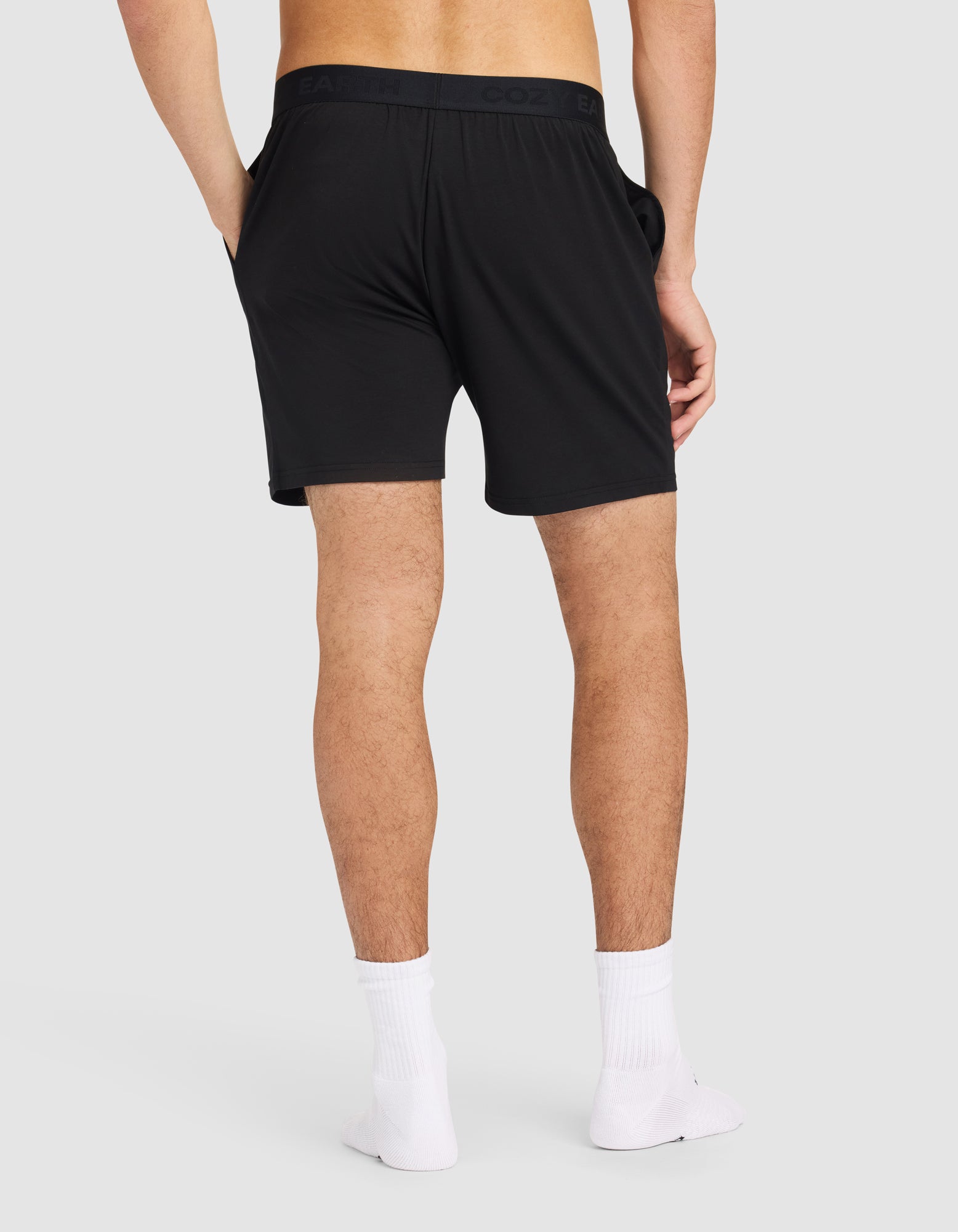A man stands with his back to the camera, hands in the pockets of Cozy Earth Men's First Layer Pajama Shorts, paired with white ankle socks and no shoes, against a plain light gray background.