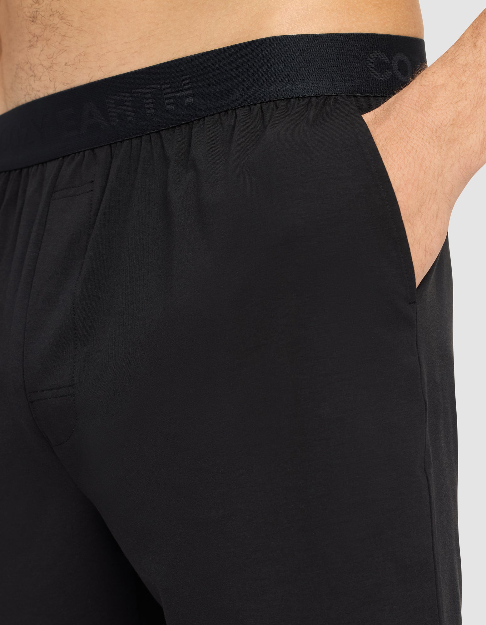 Close-up of a person wearing Cozy Earth Men's First Layer Pajama Short in black, featuring a subtle-lettered waistband and one hand in the pocket. The background is plain and light-colored.|Color:Jet Black.
