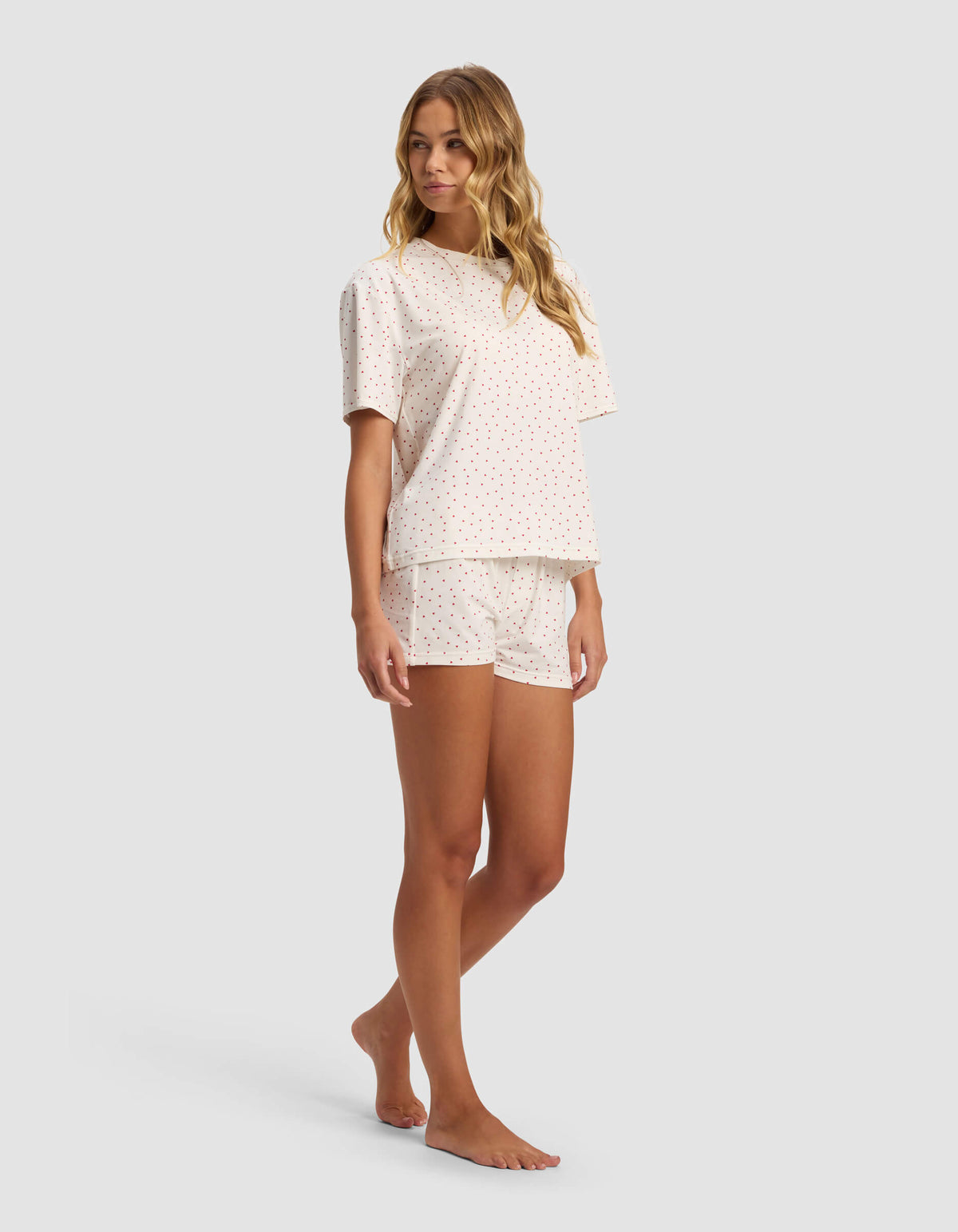 A woman stands barefoot, facing forward in a relaxed pose, wearing the Cozy Earth Women's First Layer Pajama Short Sleeve Tee & Boxer Short Set with a white base and small multicolored polka dots.