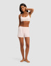 A woman stands barefoot on a light gray background, wearing the Women's First Layer Pajama Soft Bra & Boxer Short Set by Cozy Earth in pale pink. With one hand touching her head, she appears relaxed and maintains a neutral expression.|Color:Ballet
