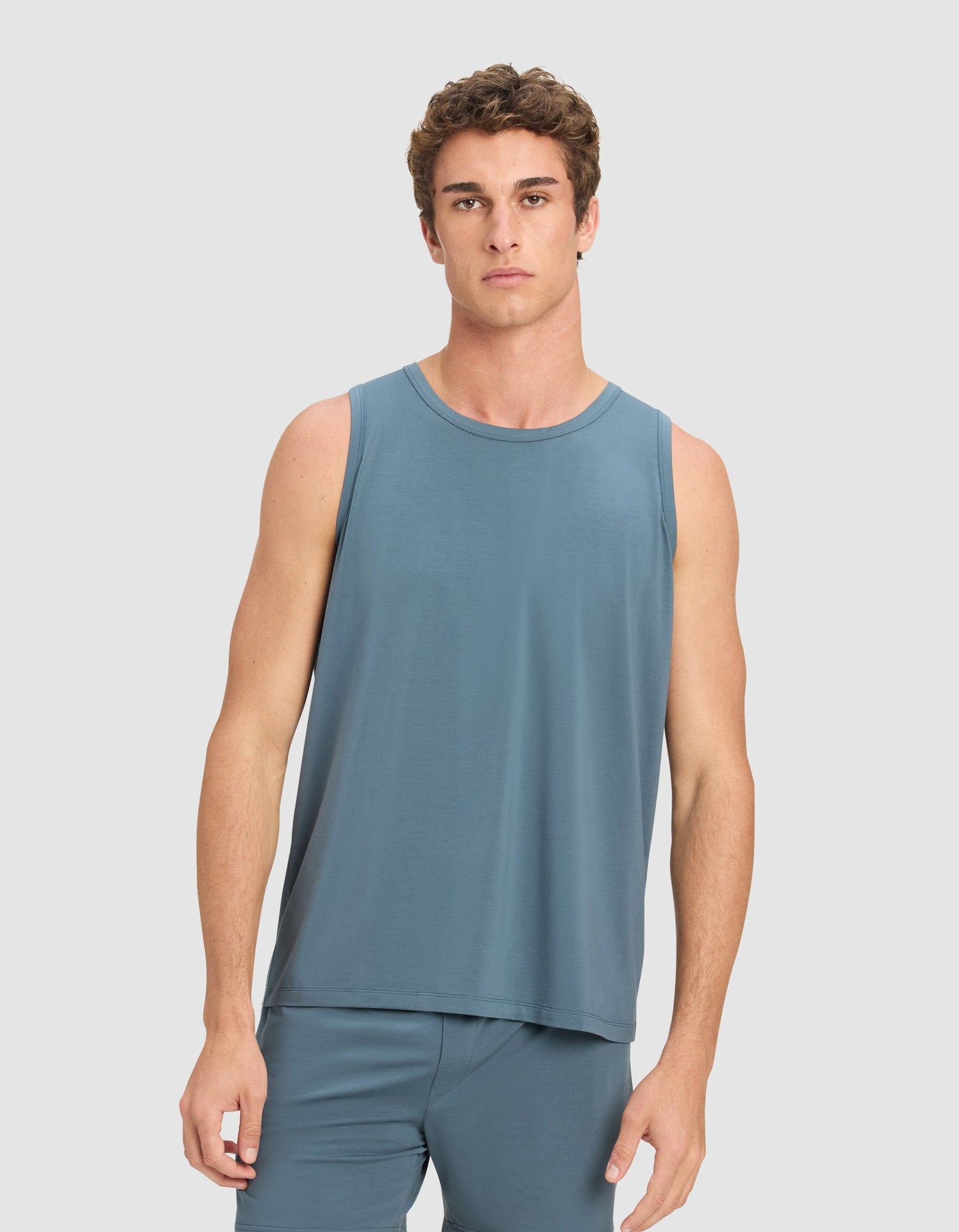 A young man with short, curly brown hair wears the Cozy Earth Men's First Layer Pajama Tank in blue, paired with matching shorts, standing against a plain light gray background and looking directly at the camera.
