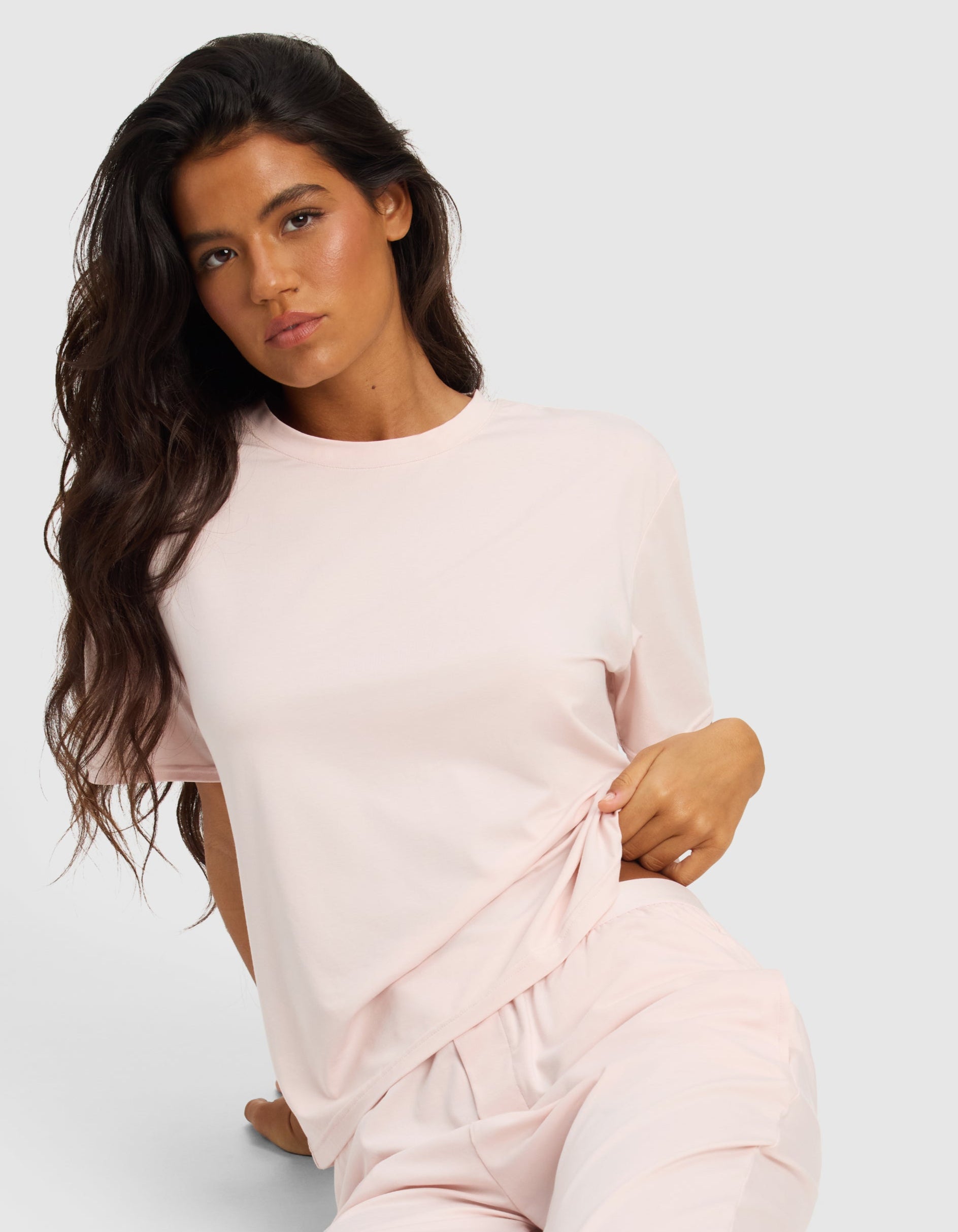 A woman with long dark hair wears the Cozy Earth Women's First Layer Pajama Short Sleeve Tee & Pant Set in light pink, sitting on the floor against a plain background and looking at the camera with a relaxed expression.