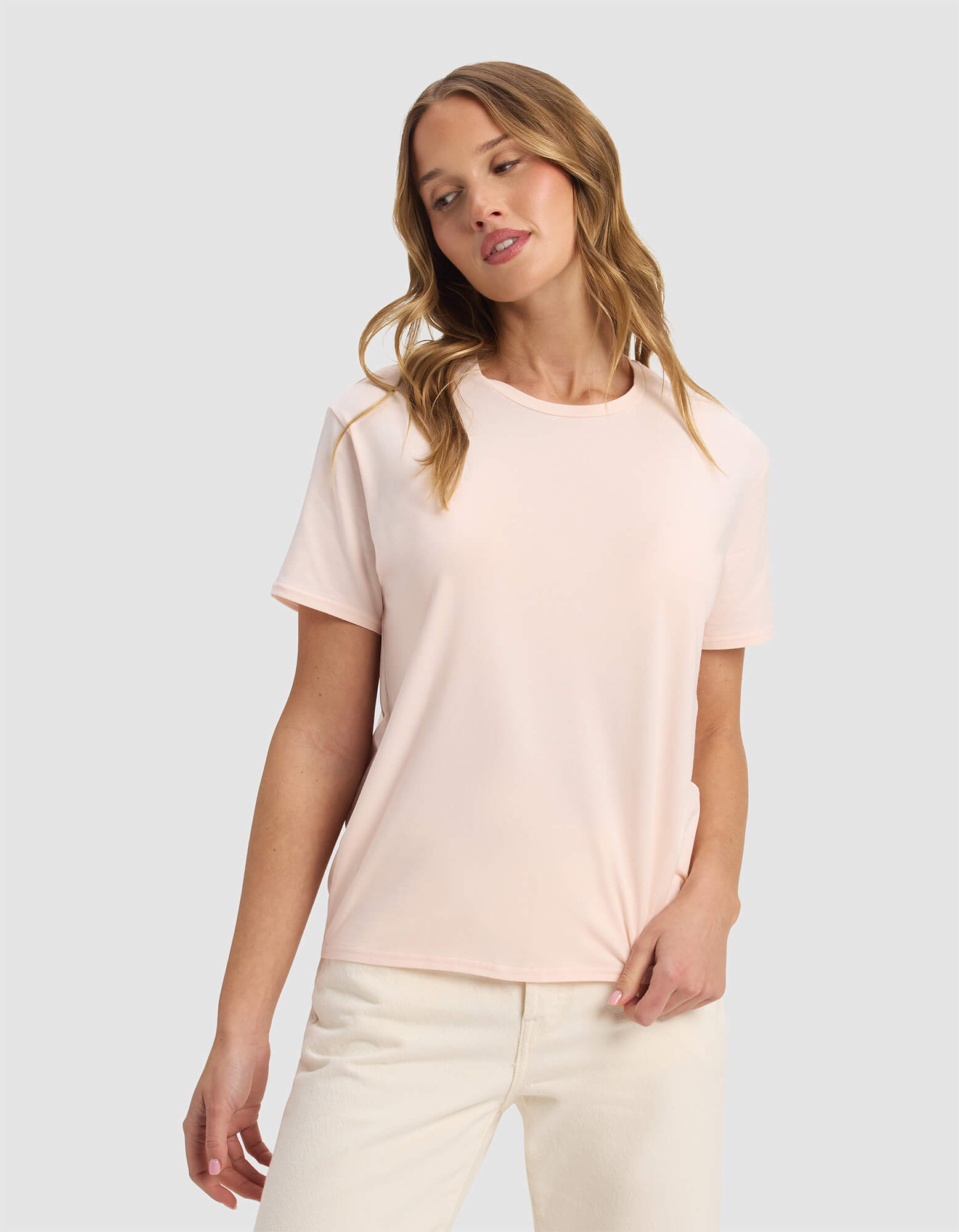 A woman with wavy blonde hair wears the Cozy Earth Women's Go-To Tee in Blossom and off-white pants, standing against a plain light gray background with a relaxed expression, looking slightly to the side.|Color:Blossom