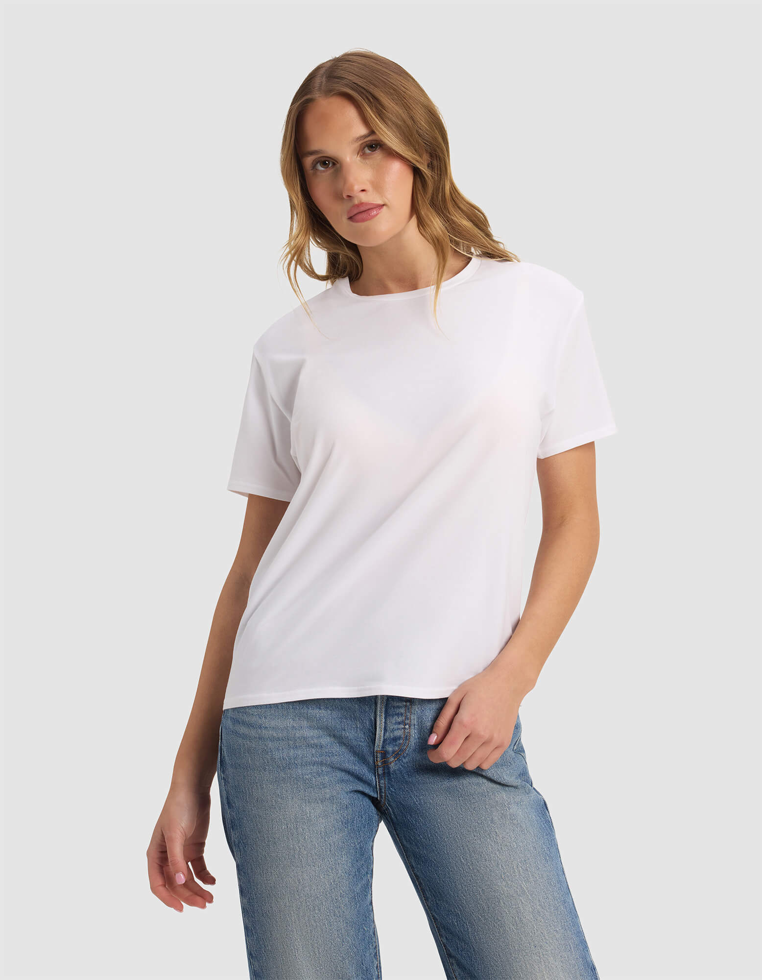 A woman with light brown hair wears the Cozy Earth Women's Go-To Tee in white and blue jeans, standing against a light gray background with one hand in her pocket and looking at the camera.|Color:White