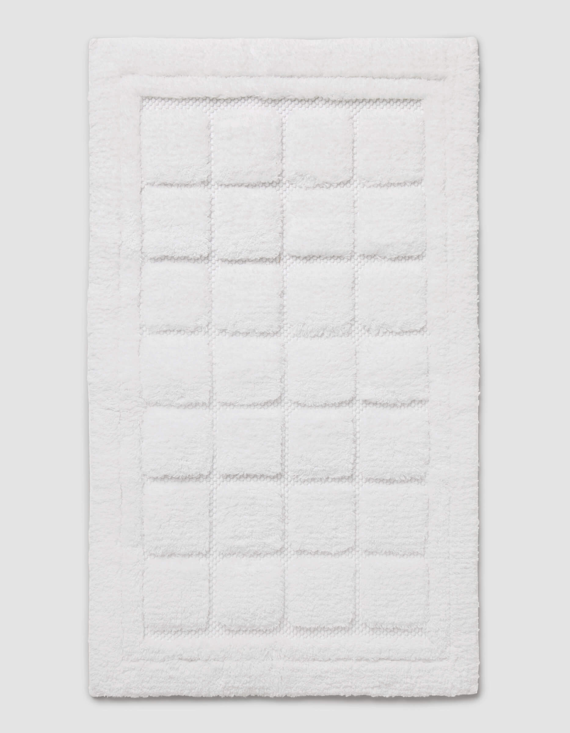 The Cozy Earth Grid Bath Mat is a white, rectangular mat featuring a textured square pattern in the center and a raised border around the edge, shown on a plain background.