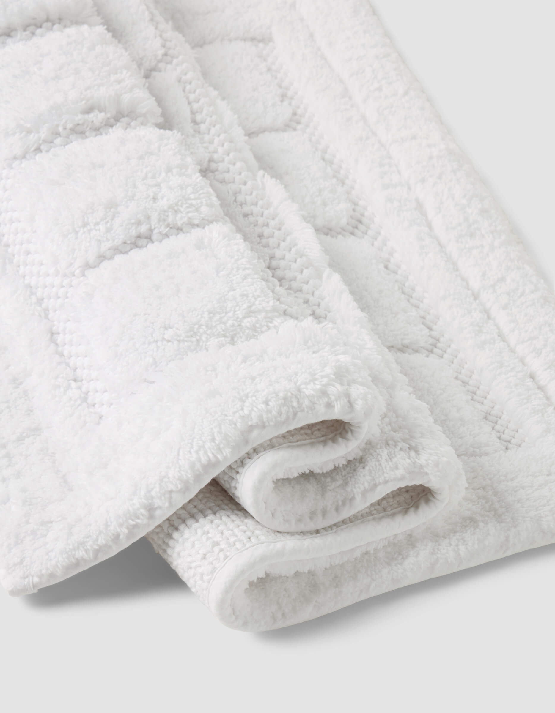 A close-up of the Folded Grid Bath Mat by Cozy Earth, featuring a white textured geometric pattern, displayed on a light gray background.