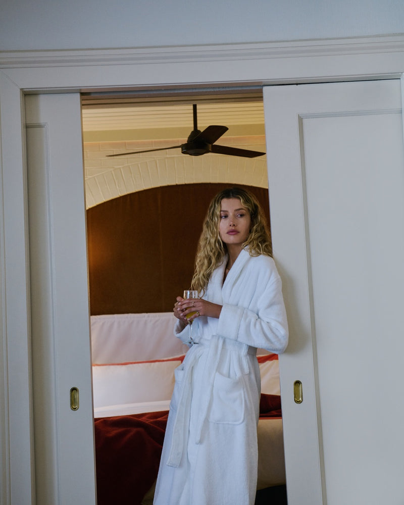 A woman with blonde curly hair wearing a white Cozy Earth Luxe Bath Robe stands in a doorway holding a glass, with a bedroom visible behind her.
