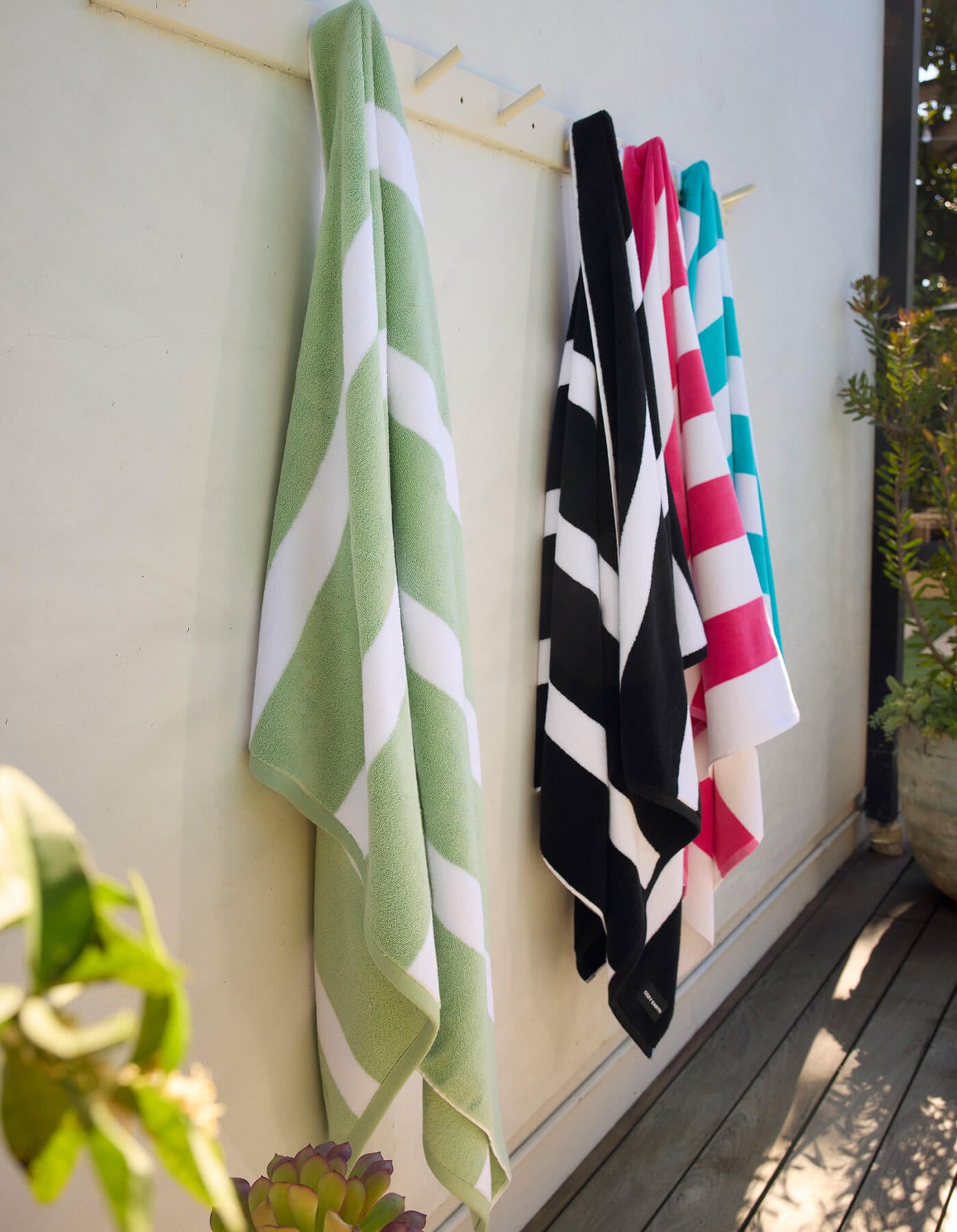 Four Cozy Earth Classic Stripe Resort Towels in green, black, red, and blue hang on hooks outside against a white wall, with plants and wooden decking nearby.