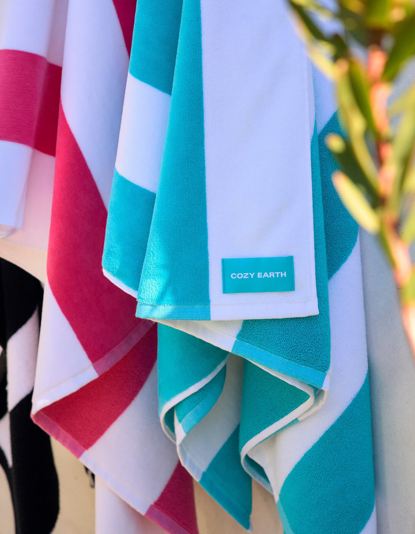 Colorful striped Seaside Resort Towels from Cozy Earth in teal, pink, and black hang side by side. The teal towel displays the Cozy Earth label. A blurred plant appears on the right.