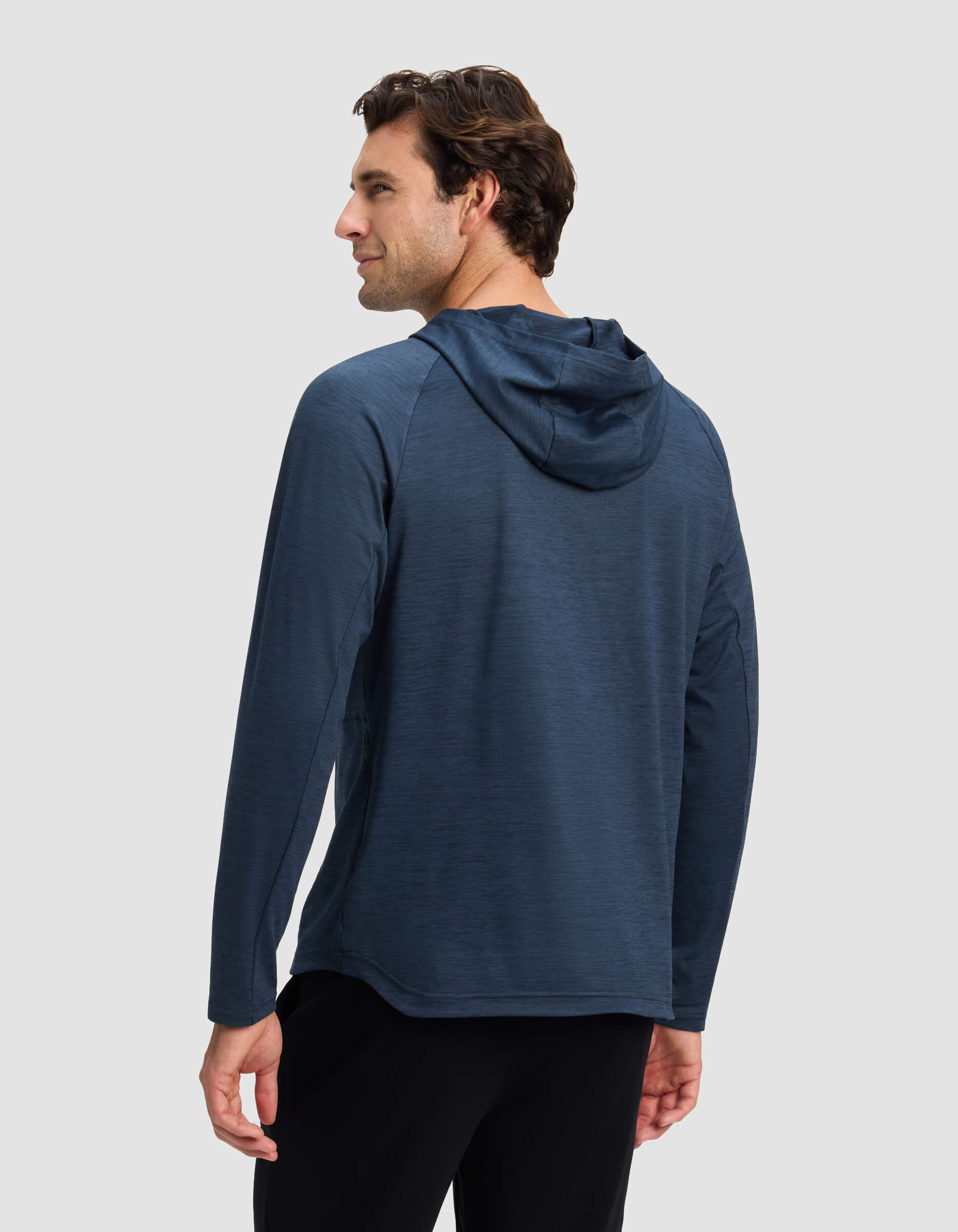 A man with short brown hair is seen from behind wearing Cozy Earth’s Men’s Lightweight Performance Hoodie in dark blue and black pants, standing against a plain light gray background.