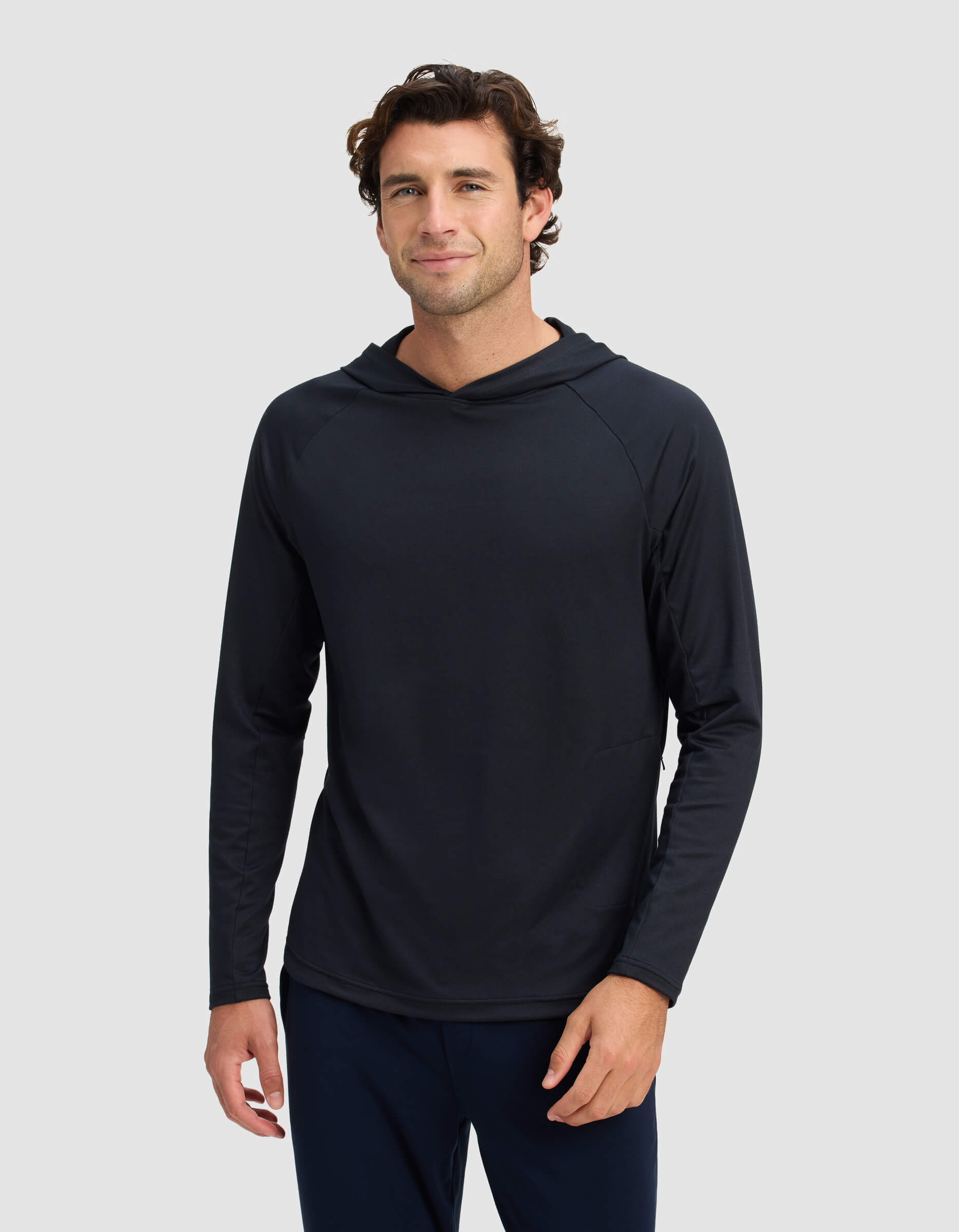 A man with short, wavy brown hair is wearing the Men's Lightweight Performance Hoodie by Cozy Earth, standing against a plain light gray background and smiling slightly at the camera.