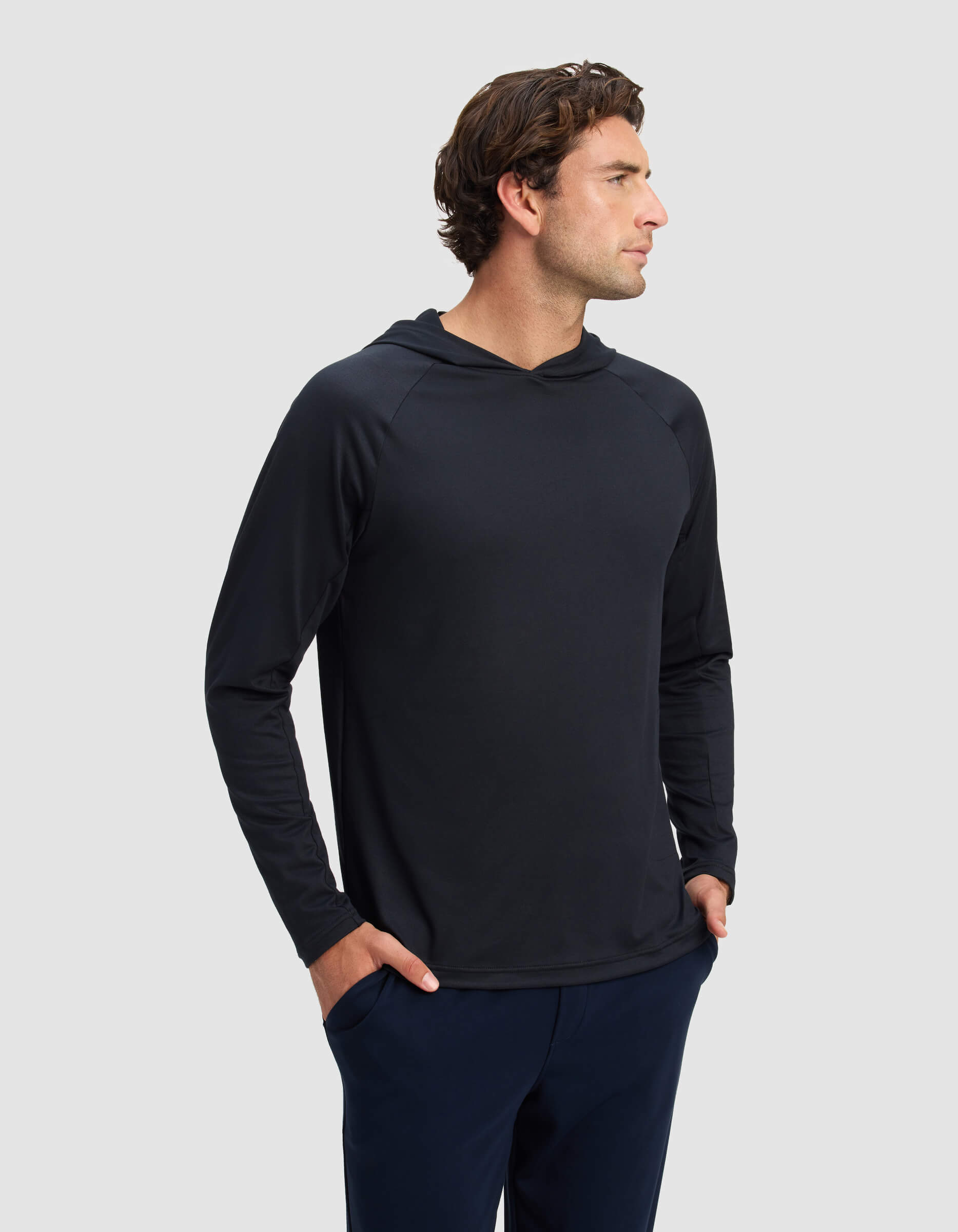 A man with wavy brown hair, wearing the Cozy Earth Men's Lightweight Performance Hoodie and dark pants, stands with his hands in his pockets, looking to the side against a plain light gray background.
