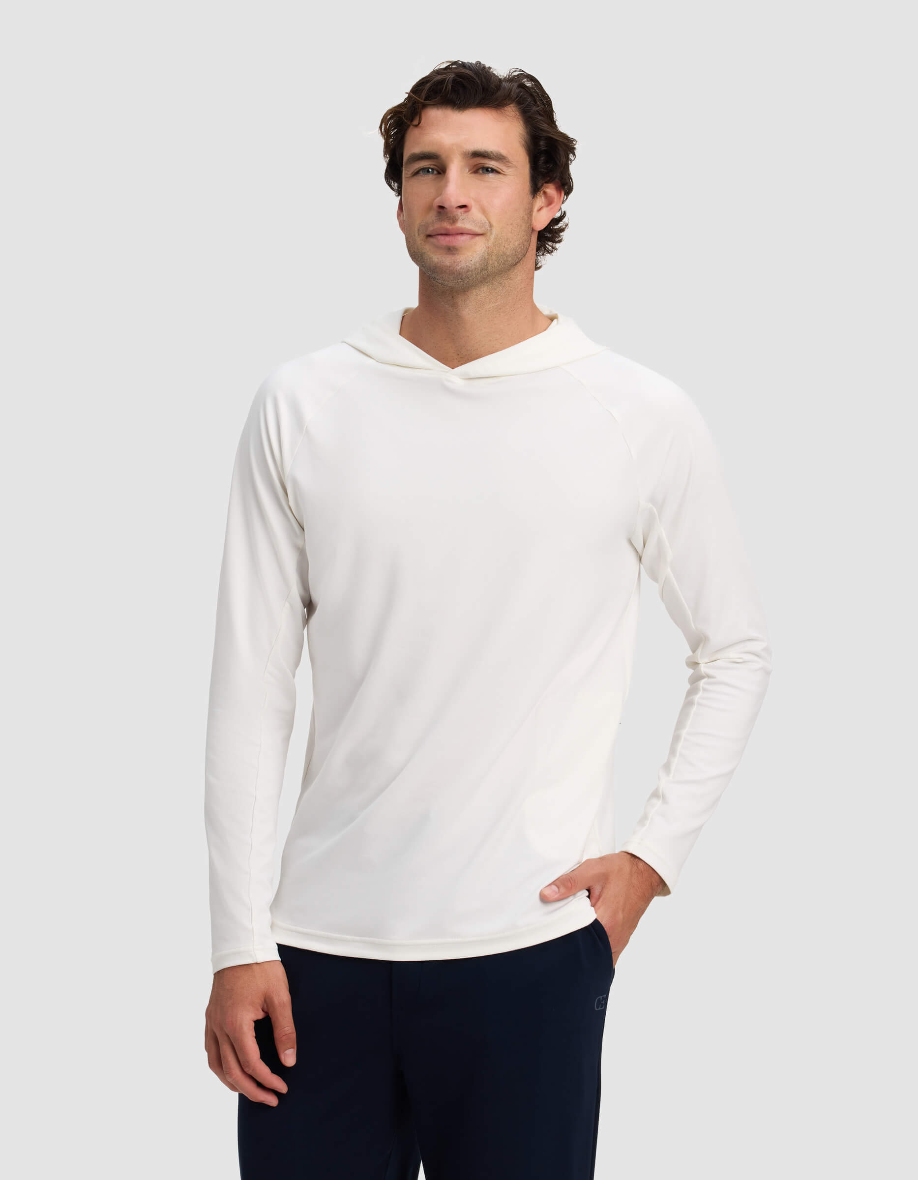 A man wears the Cozy Earth Men's Lightweight Performance Hoodie in white with dark pants, standing against a light gray background, facing forward with one hand in his pocket and a slight smile.|Color:White