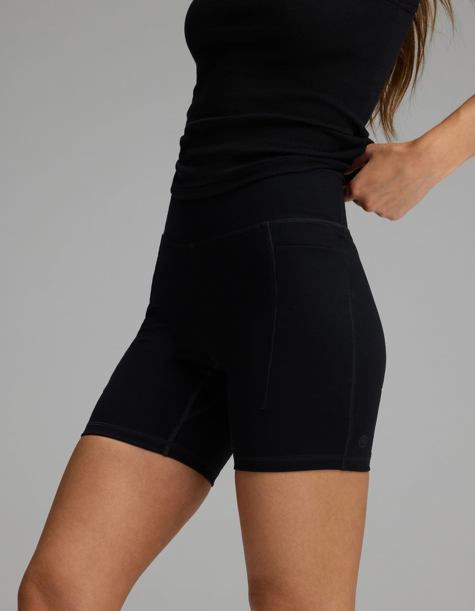 A person wearing black Women’s Liv Biker Short 6″ with Pockets, featuring a high-waisted fit and side pockets.|Color:Jet Black