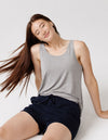 A young woman with long brown hair sits on the floor, smiling with her eyes closed, wearing the Cozy Earth Women's Bamboo Stretch-Knit Lounge Tank in grey, paired with navy shorts against a plain white background.|Color:Grey