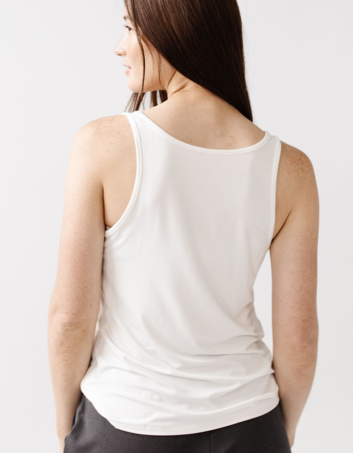 A woman with long brown hair is seen from behind wearing the Cozy Earth Women's Bamboo Stretch-Knit Lounge Tank in white, paired with dark pants, standing before a plain light background.|Color:Ivory