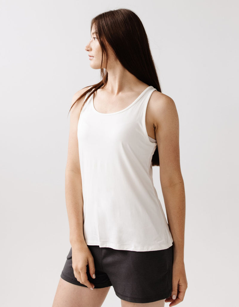 A woman with long brown hair wears the Cozy Earth Women's Bamboo Stretch-Knit Lounge Tank in Ivory, paired with dark shorts, standing against a plain light background and looking to her left.
