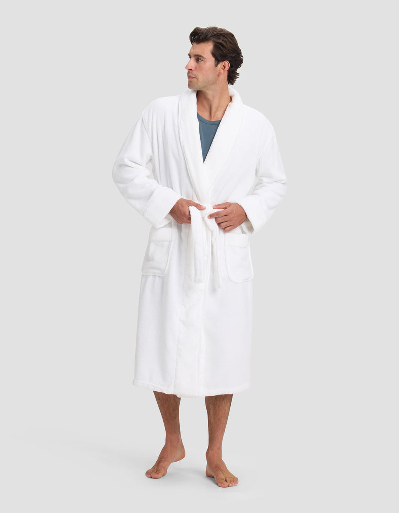 A man with short, dark hair stands barefoot in a Cozy Earth Luxe Bath Robe over a blue shirt, tying the belt at his waist. He faces slightly right against a plain light gray background.