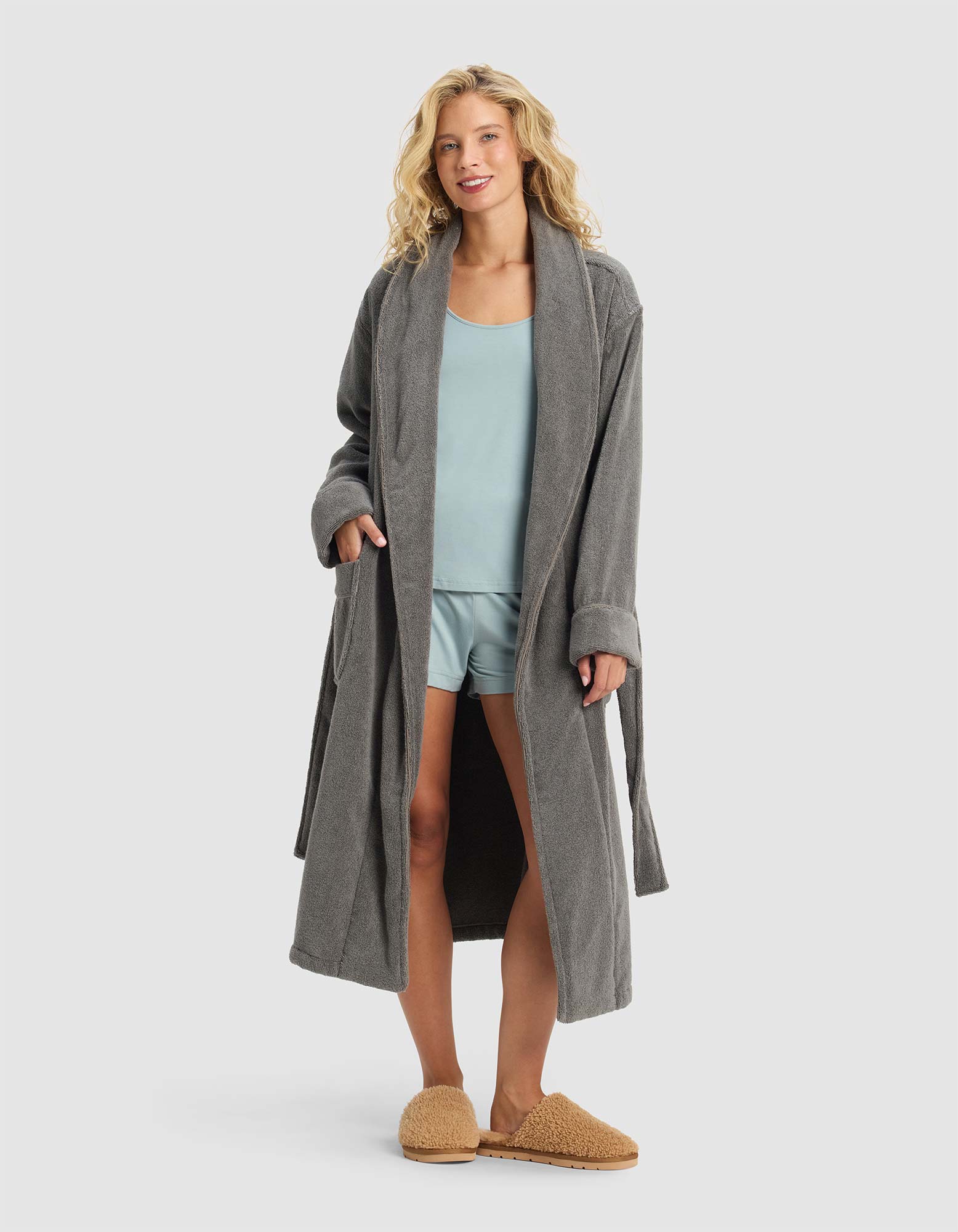 A woman smiles with her hands in the pockets of a Luxe Bath Robe by Cozy Earth, layering it over light blue pajamas and tan slippers against a plain light gray background.