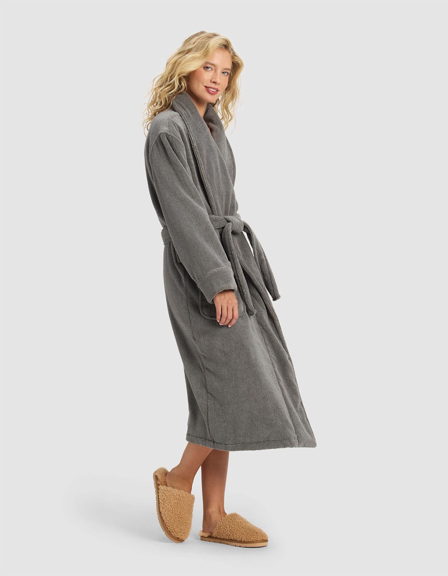 A person stands sideways in the Cozy Earth Luxe Bath Robe, a long gray robe with a waist belt, paired with tan fuzzy slippers against a plain light background.