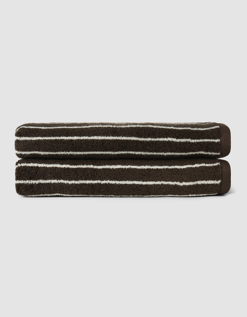 The Complete Striped Luxe Bath Bundle by Cozy Earth features two dark brown towels with thin, irregular white stripes, neatly rolled and stacked on a light gray background.