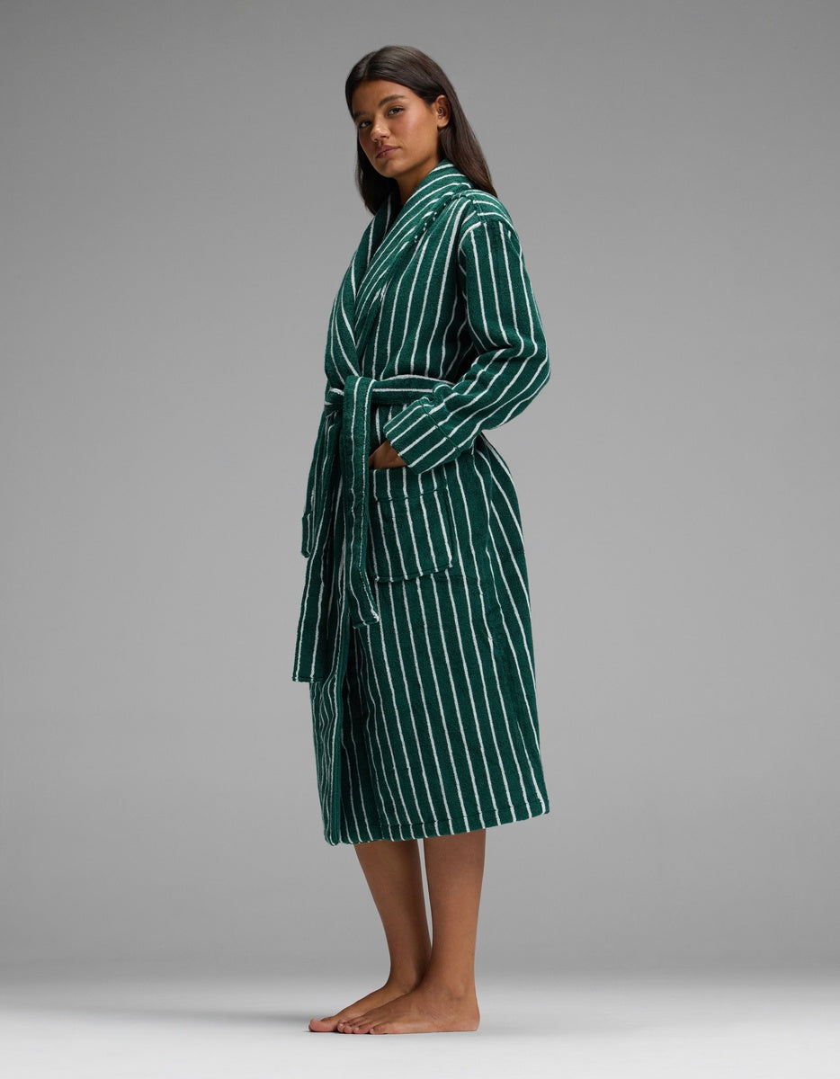 A woman stands barefoot in the Luxe Bath Robe by Cozy Earth, featuring green fabric with white vertical stripes, hands in the pockets, against a plain gray background.